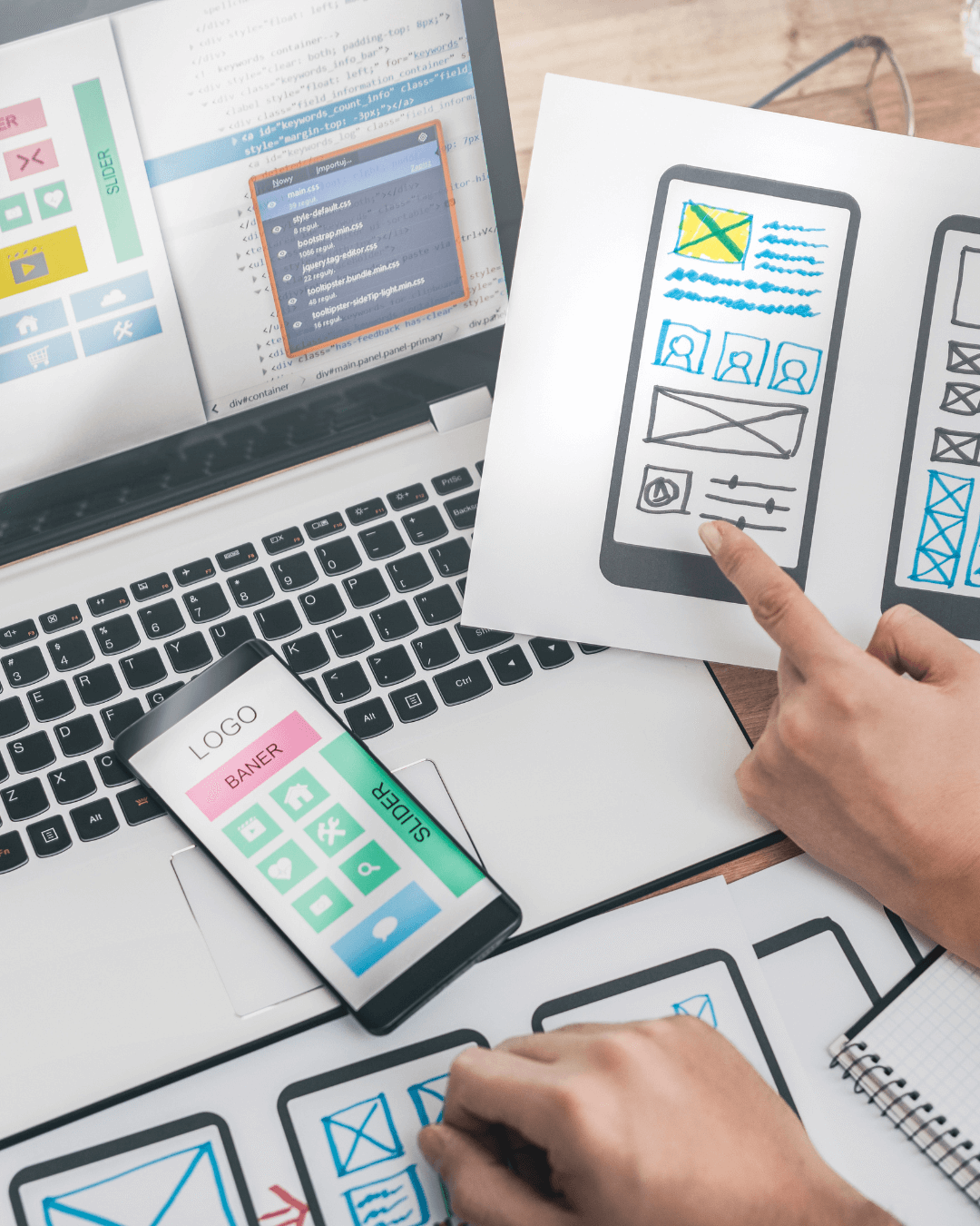 Person zeigt auf ein gezeichnetes Smartphone-Layout – Webdesign-Konzept liegt auf Laptop mit offenem Code-Editor